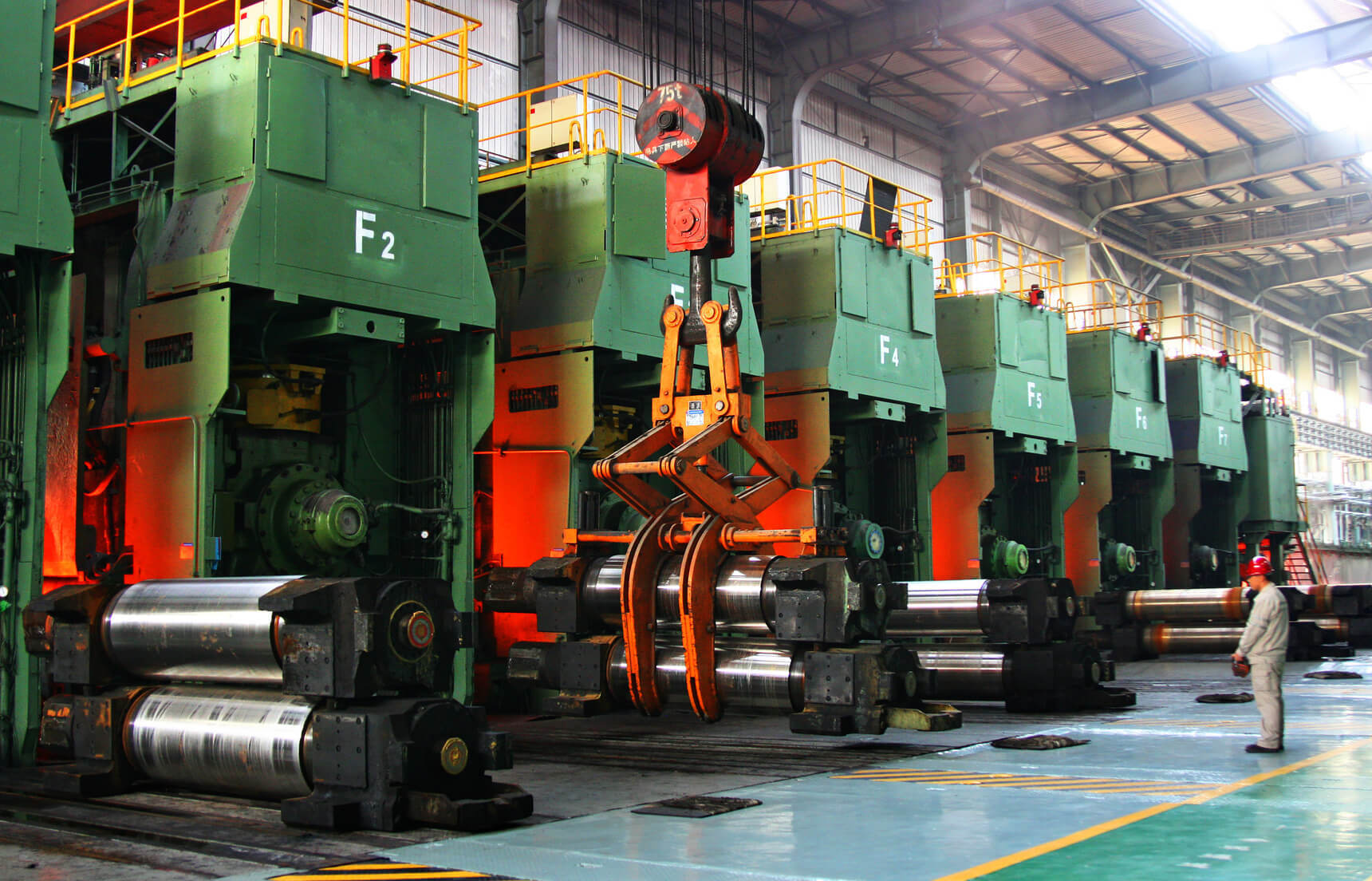 A factory with large green machinery labeled "F2" along one side, showcasing heavy industrial equipment. An orange robotic arm is positioned in the middle, gripping metal sheets. A worker in a helmet stands on the right side of the image, observing the operations.